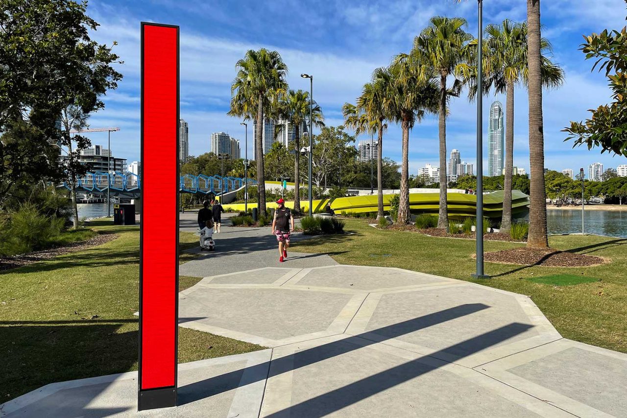 A slim red digital column among palms and lawns at the HOTA cultural precinct.