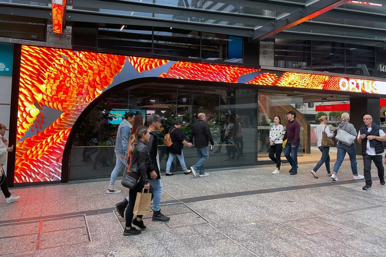 Pedestrians pass the Optus store beneath a vivid red and yellow digital display.