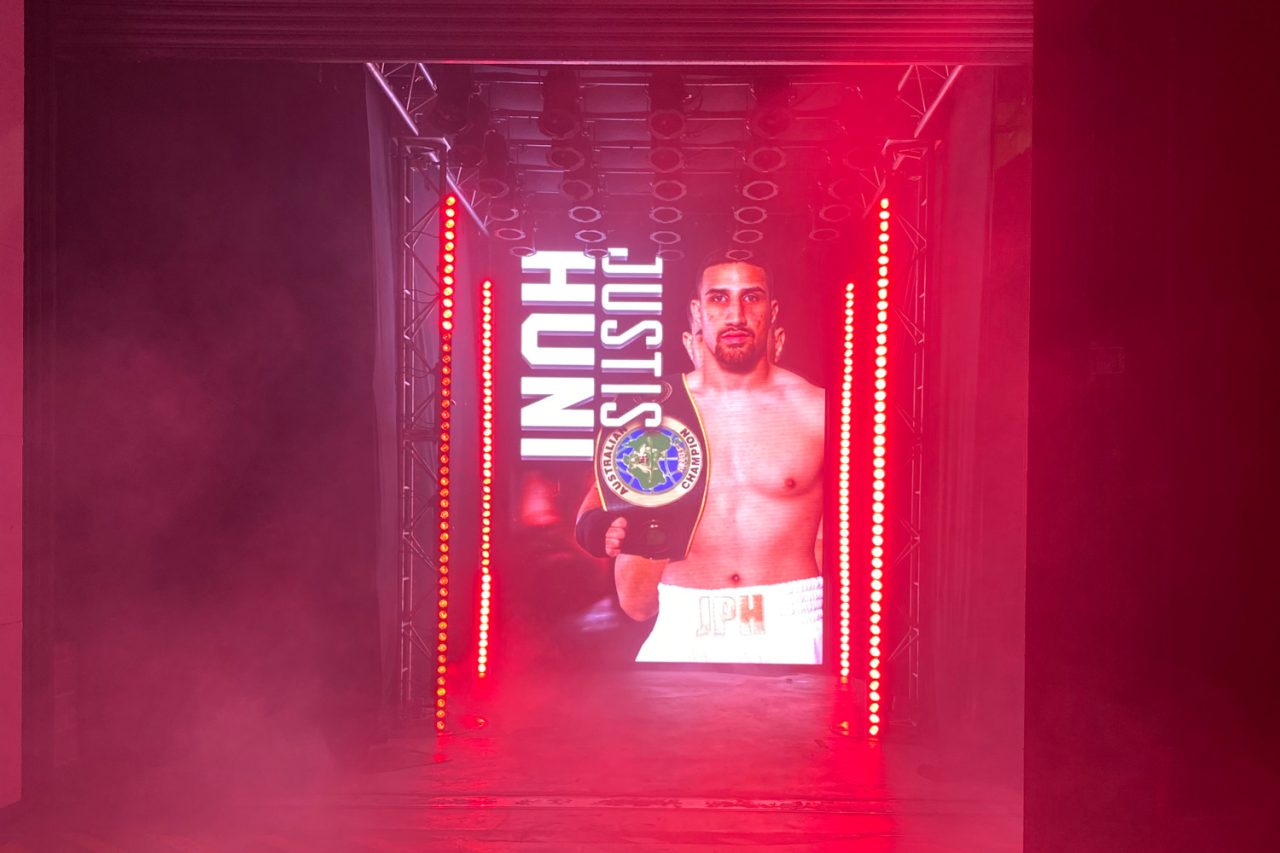 Justis Huni graphic on a tall entrance screen amid red light and haze.