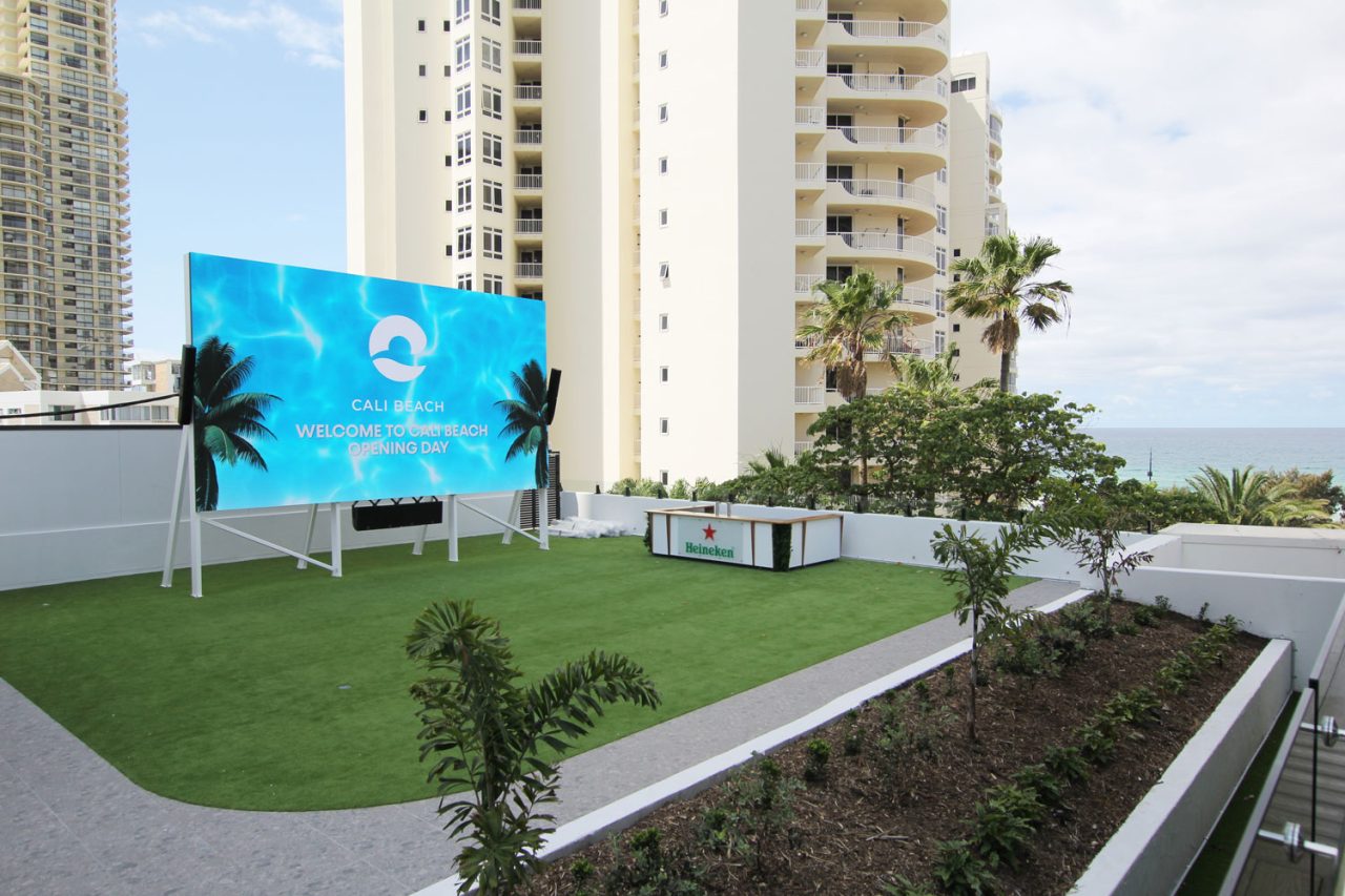 Outdoor screen beside the lawn at Cali Beach, with Gold Coast towers and the ocean behind it.