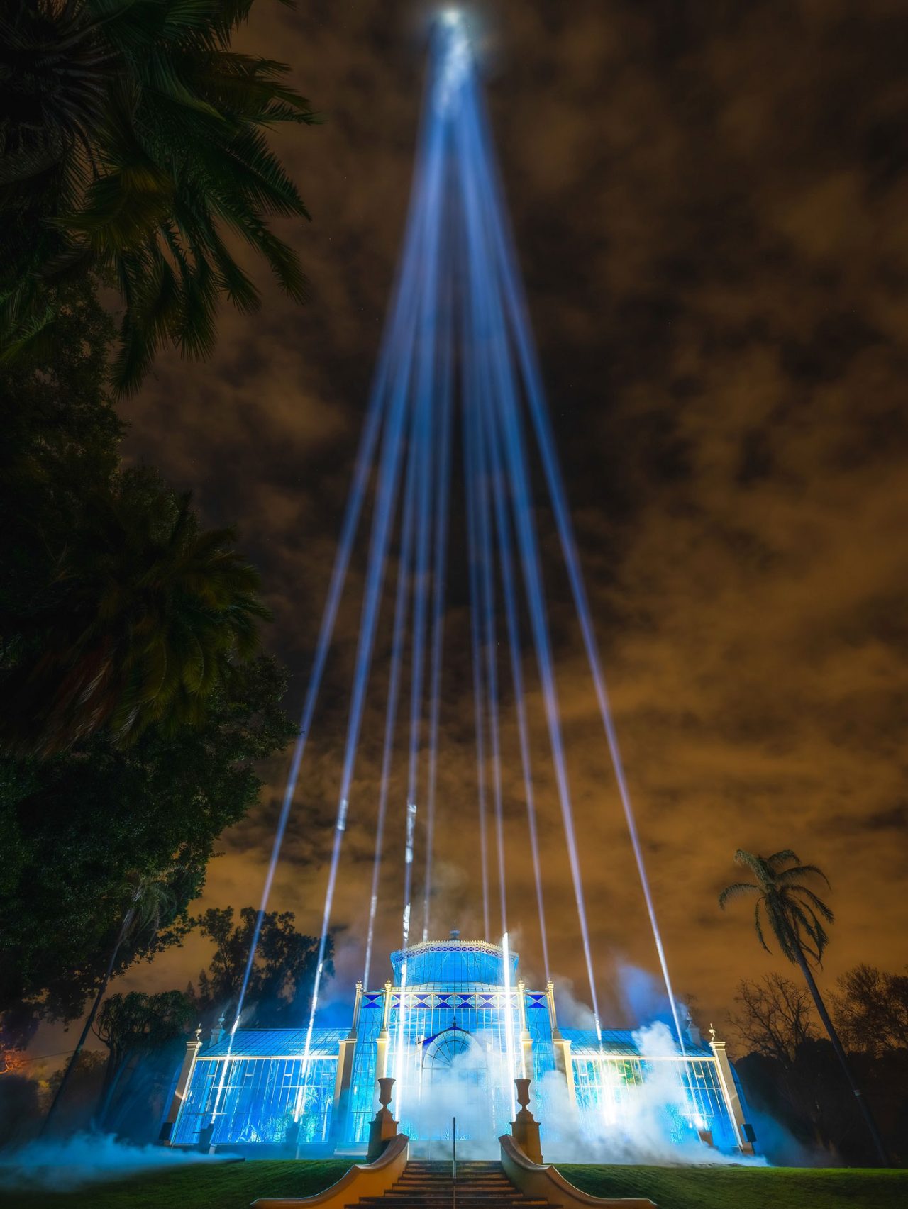 White beams converge above a blue-lit glasshouse at Illuminate Adelaide's Light Cycles.