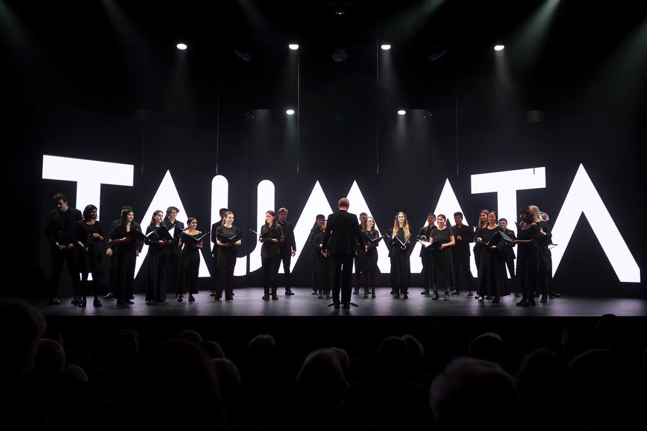 Performers silhouetted against large illuminated Taumata letters.