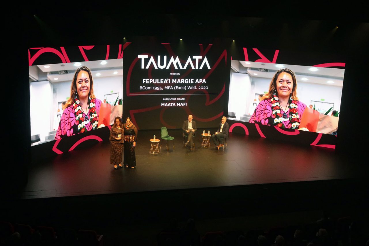 A speaker on stage between large portrait screens at the University of Auckland's Taumata 2022 event.