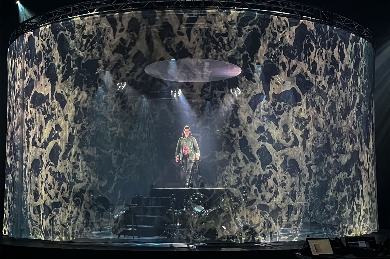 Performer inside a circular set surrounded by textured imagery and light.
