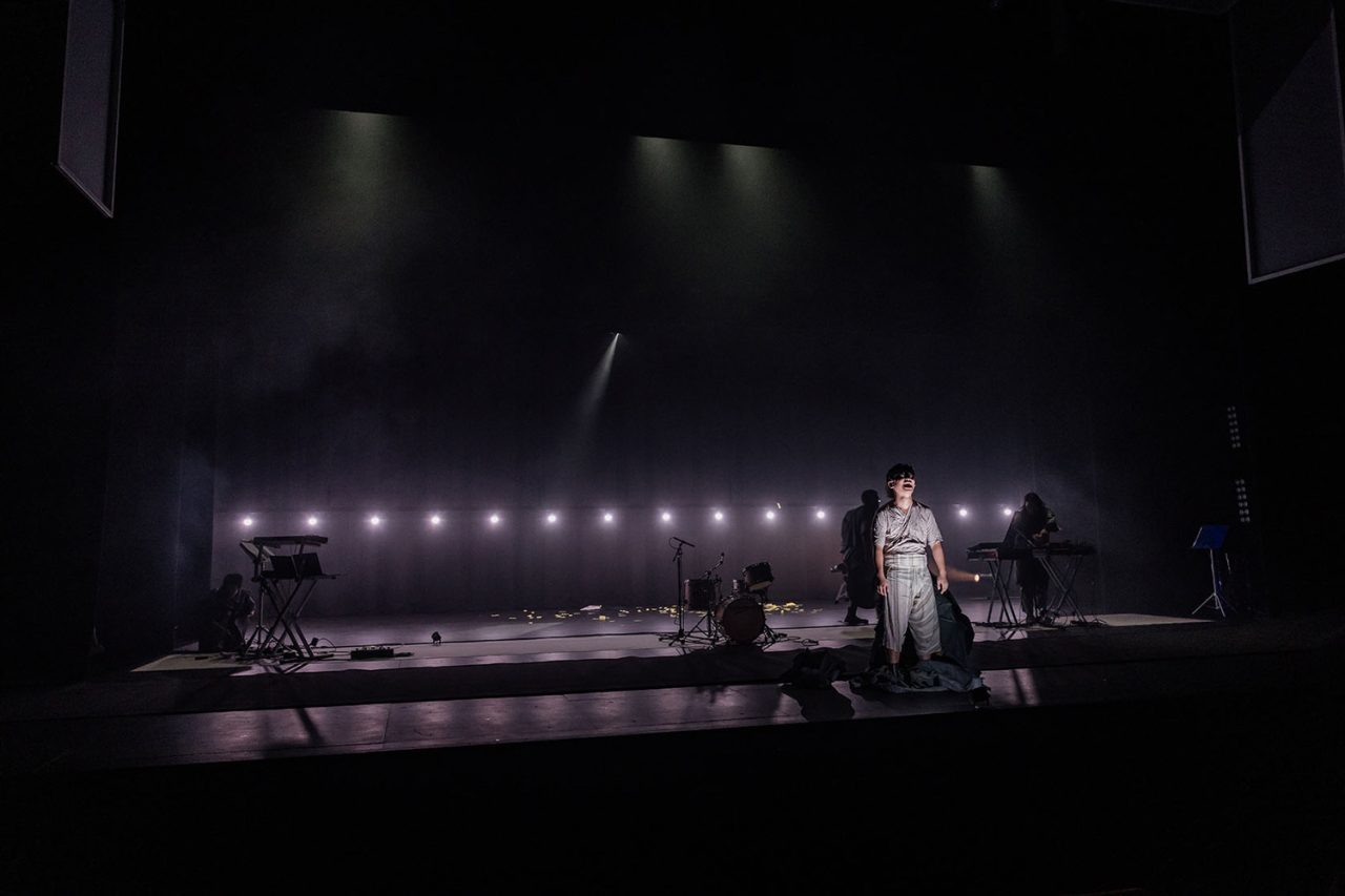 Performer on a dark stage beside a horizontal row of small lights.
