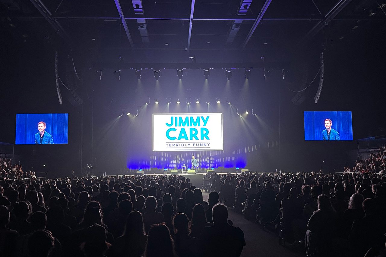 Audience facing the central Terribly Funny screen and two side screens at the Jimmy Carr show.