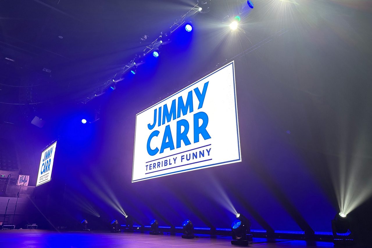 Jimmy Carr Terribly Funny branding on the central screen beneath blue stage lighting.