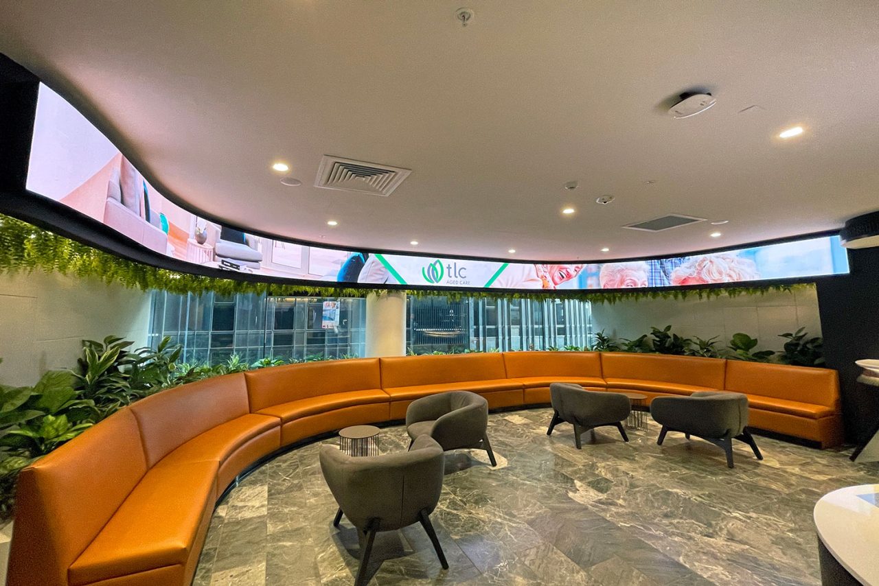 Curved orange seating beneath the wraparound digital ribbon at TLC Aged Care reception.