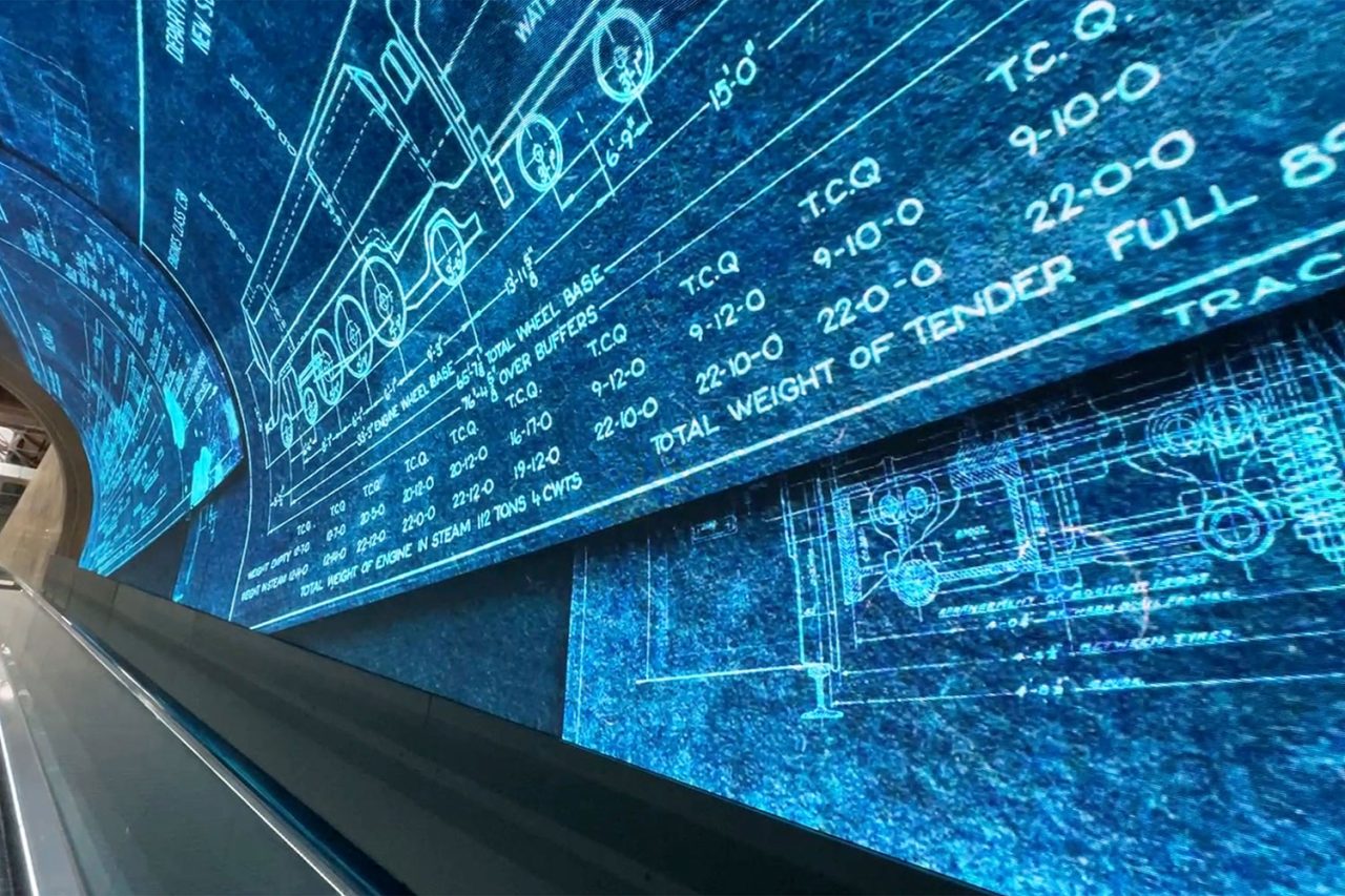 Blue technical drawings fill the curved digital display beside the Foundry Tunnel escalator.