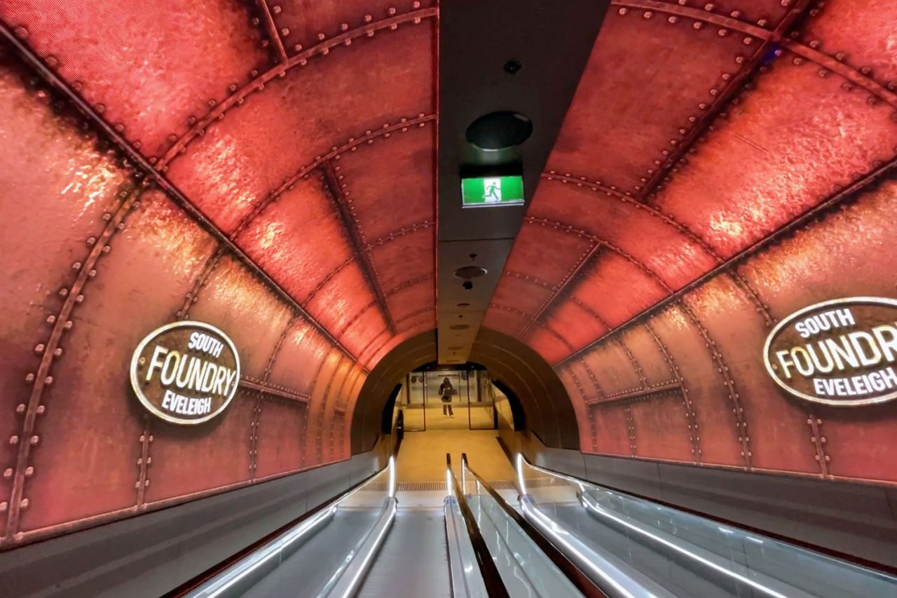 Curved LED walls surround the escalators in South Eveleigh’s Foundry Tunnel, displaying red industrial-themed graphics.