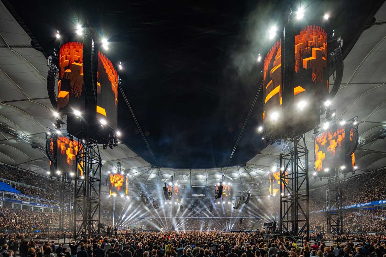 Illuminated video towers and white beams above the M72 stadium audience at night.
