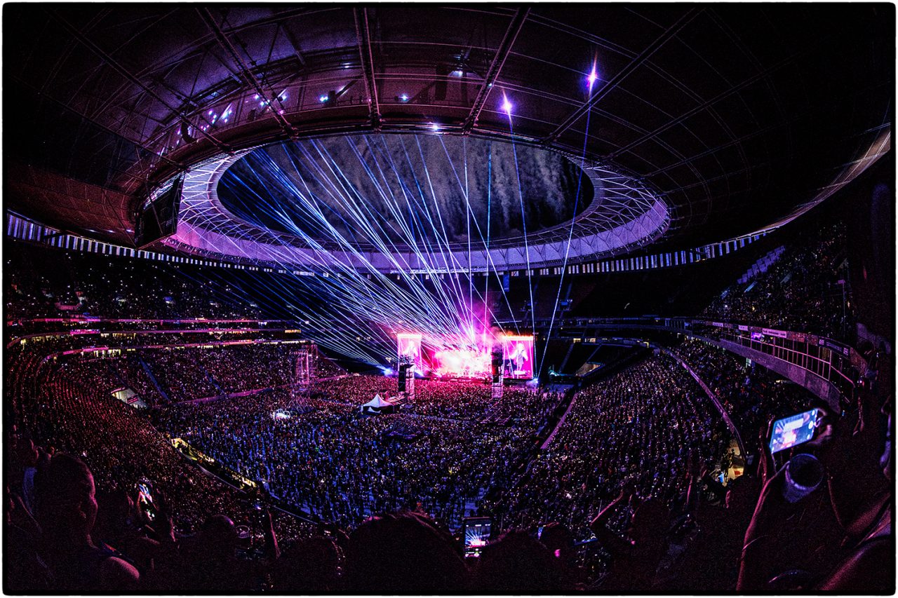 Packed stadium with white beams extending from the central stage beneath a circular roof.