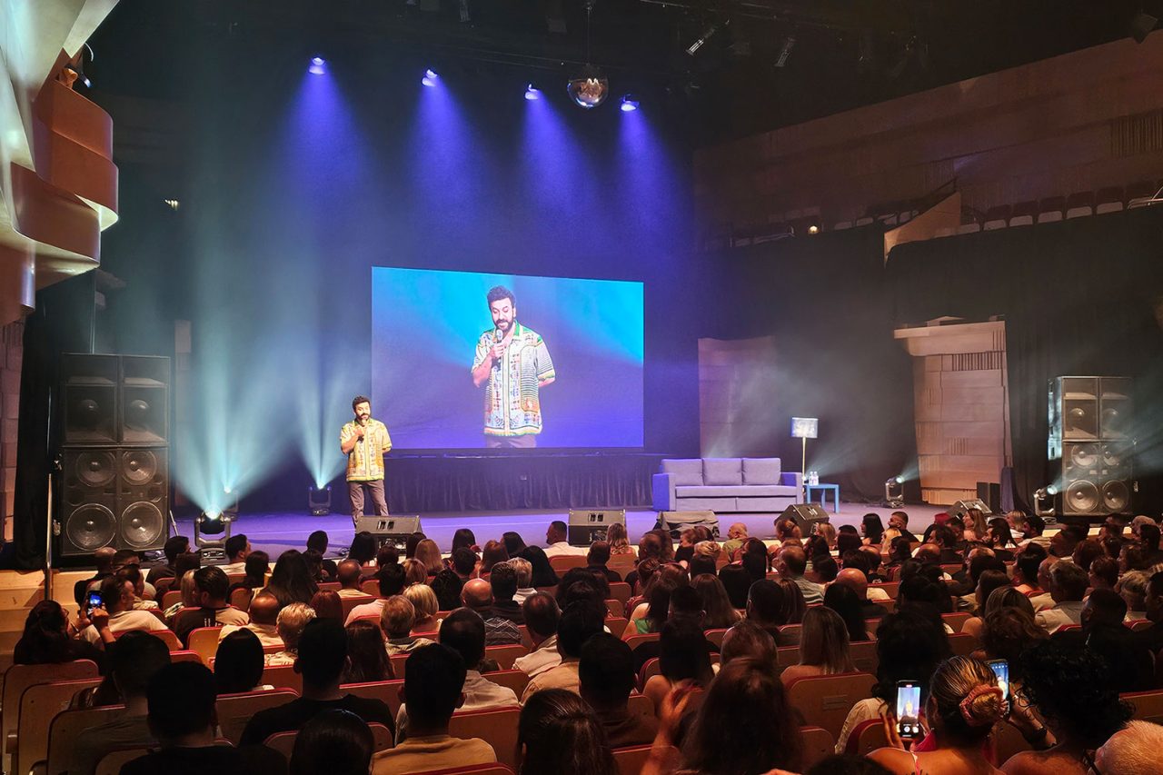 Speaker onstage with a live camera feed displayed on a large screen above the audience.