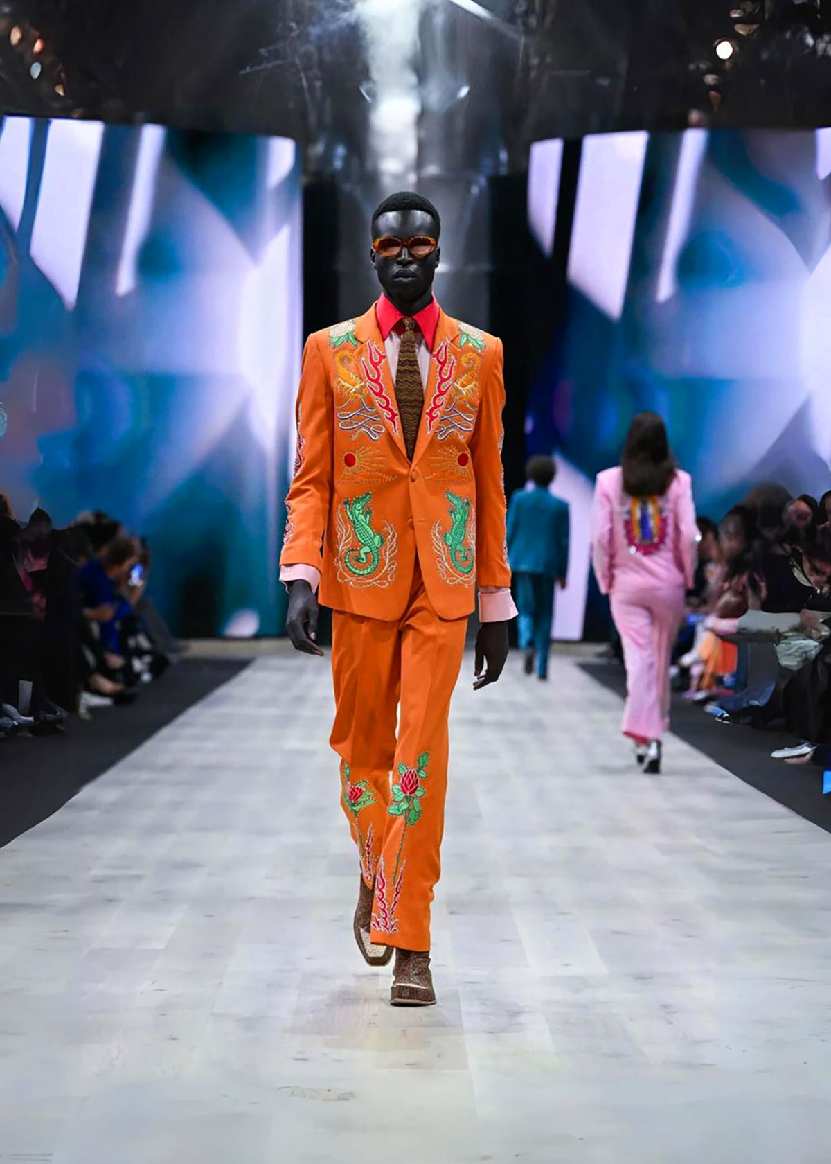 Model in an orange outfit walking the runway, with another model dressed in pink behind.