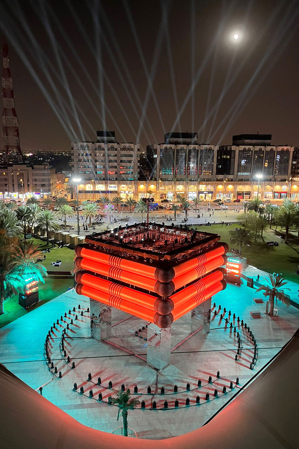 Aerial view of a red cube-like installation in a turquoise-lit courtyard.
