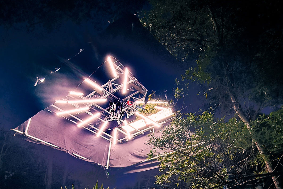 Overhead view of an outdoor lighting rig with bright LED tubes among trees at night.