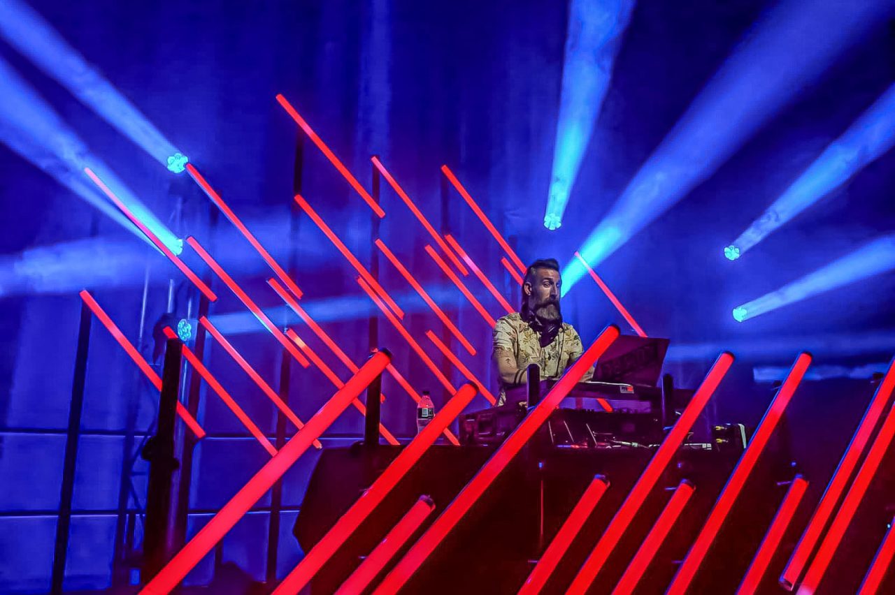 DJ framed by diagonal red LED tubes and blue beams.