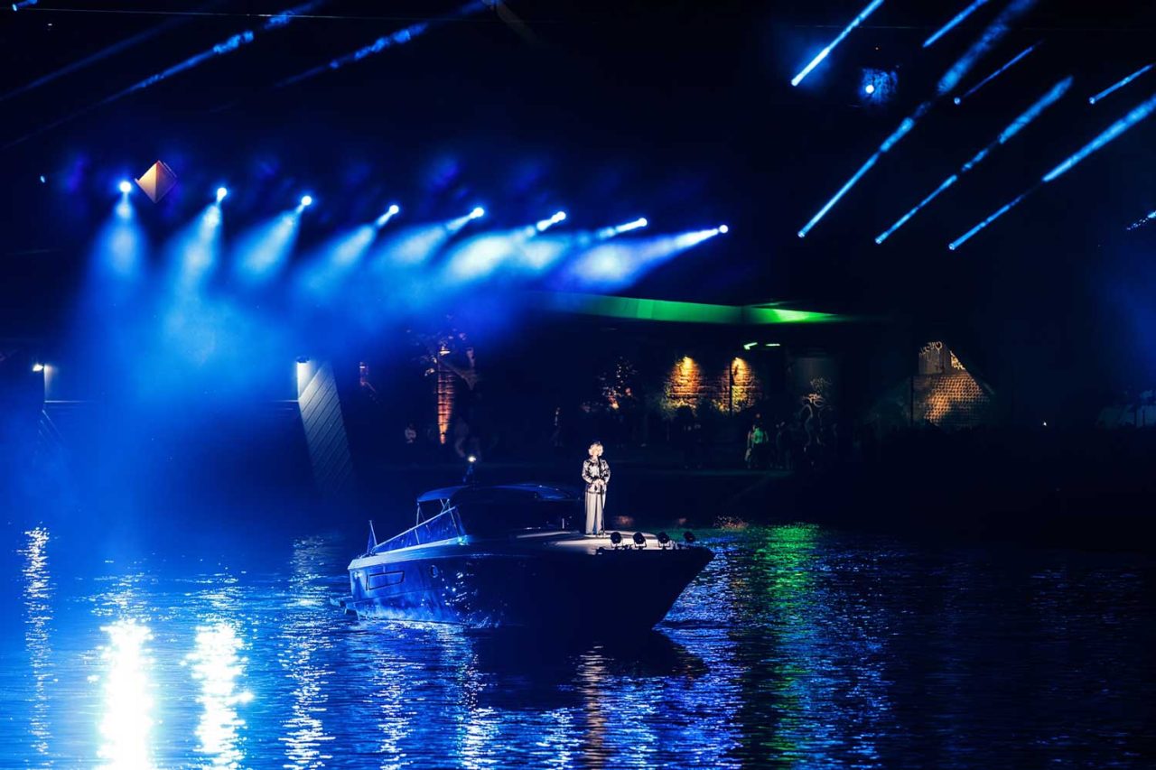 Performer on a platform over the river beneath blue and white stage lighting.