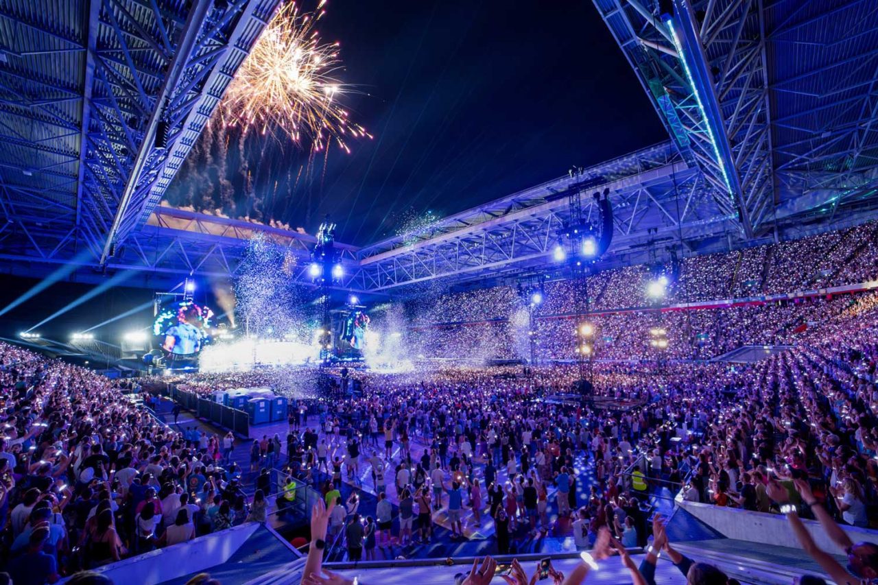 Fireworks above a crowd illuminated in blue during Coldplay's stadium show.