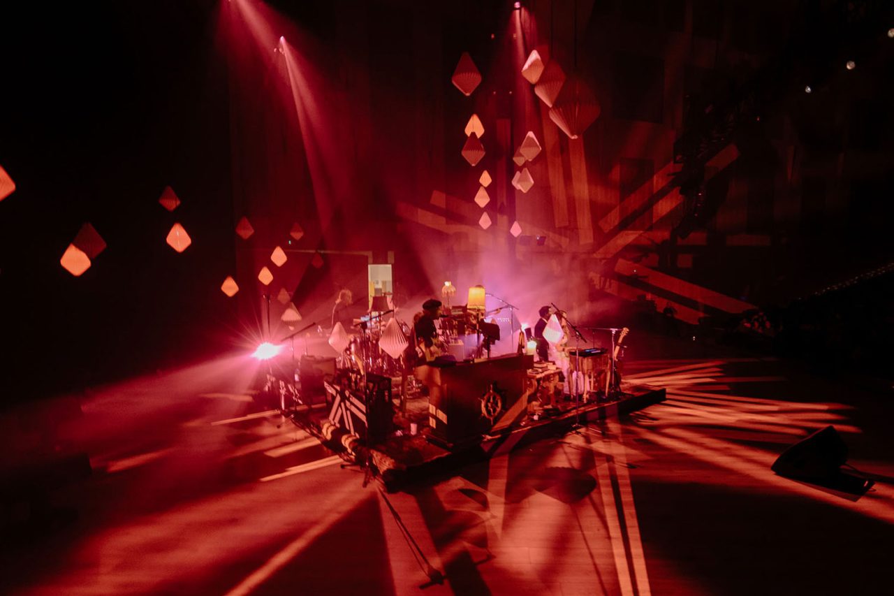 Band performing among hanging lamps under red light on the Living Room Sessions tour.