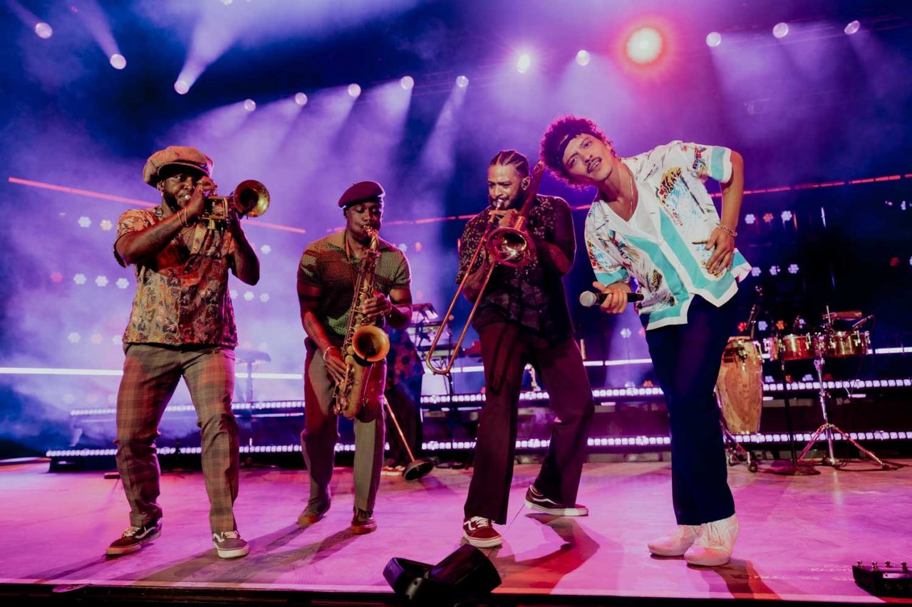 Musicians performing together under purple and pink stage lights at a Bruno Mars concert.