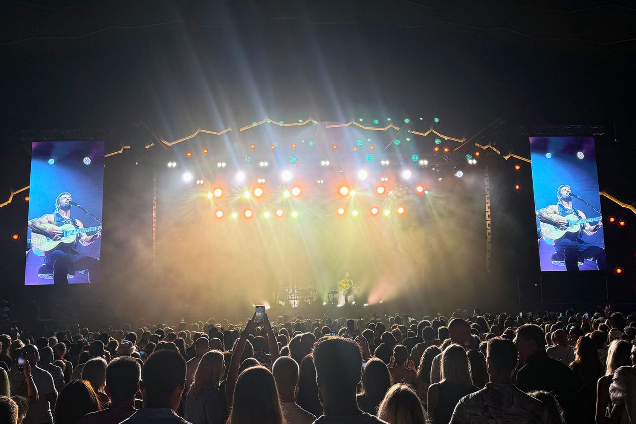 Audience facing warm stage lighting and side screens during Xavier Rudd's Freedom Sessions show.