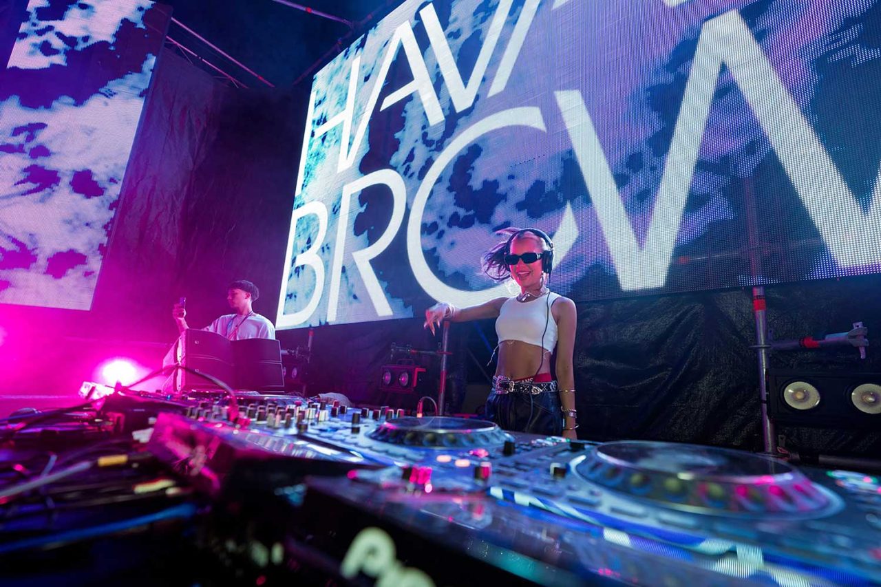 DJ performing behind mixing decks with Havana Brown graphics on the screen.