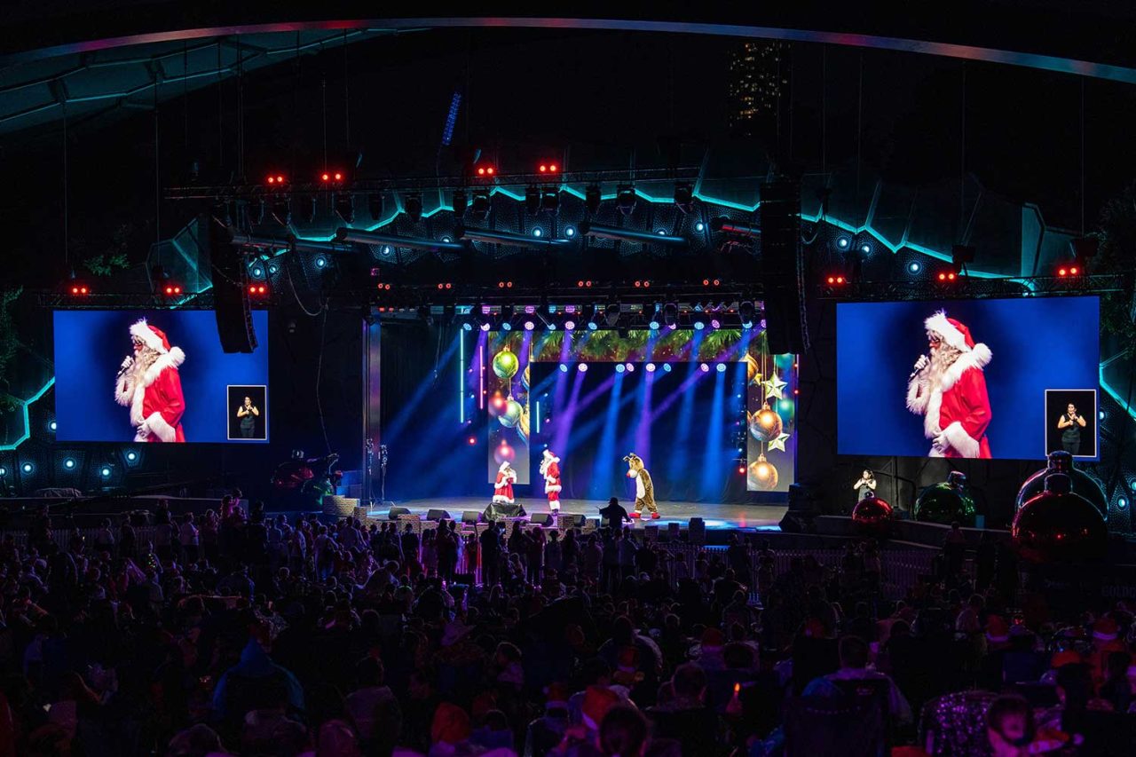 Performer in red shown on both side screens above the carols audience.
