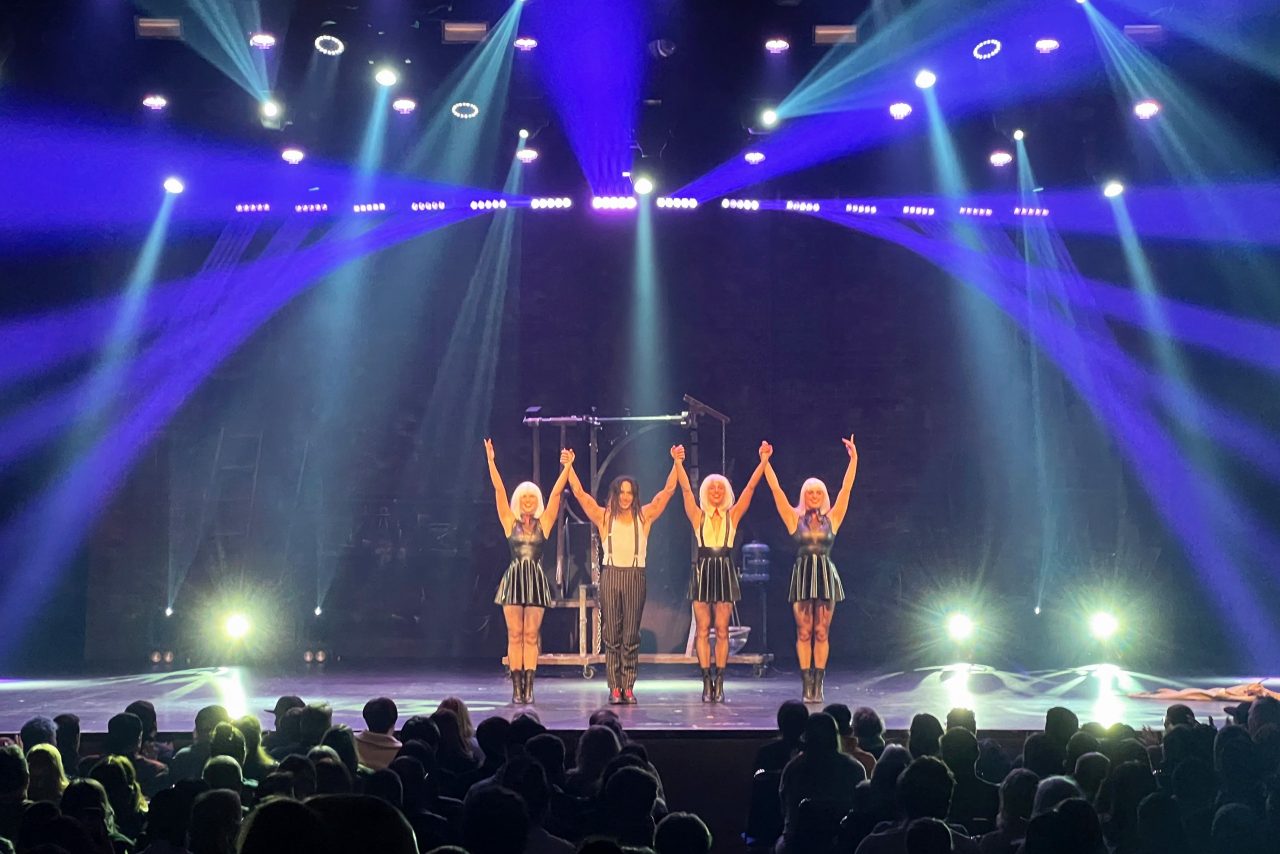 Performers raise their arms beneath blue and white beams at the end of the Decennium show.