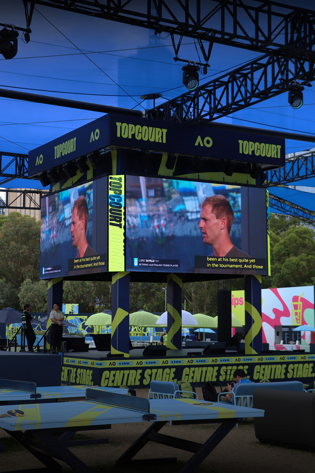 Large suspended video screens above tables in the Australian Open Topcourt fan zone.