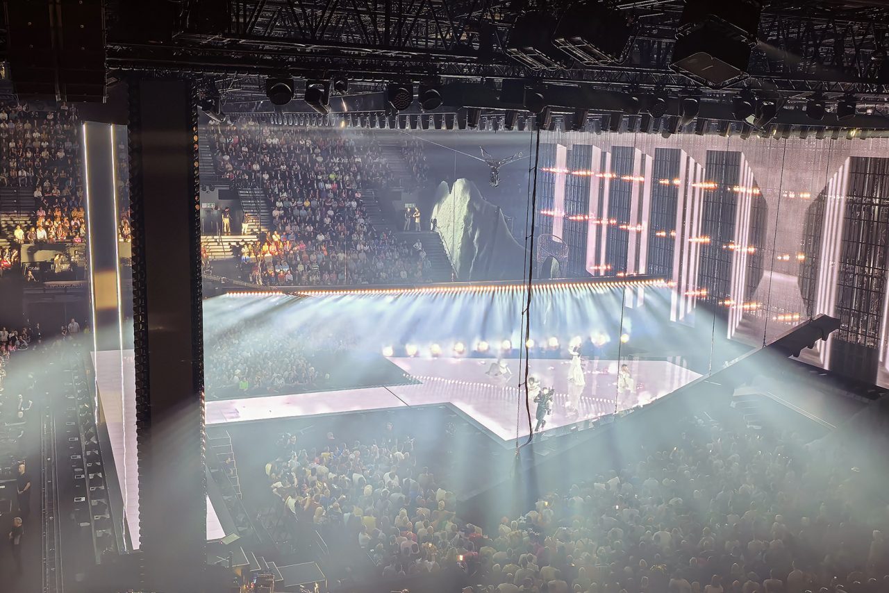White beams passing through haze above the Eurovision audience and stage.