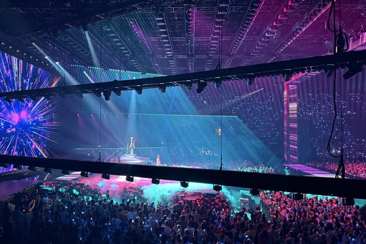 Overhead lighting rig above blue stage screens and a pink-lit Eurovision crowd.