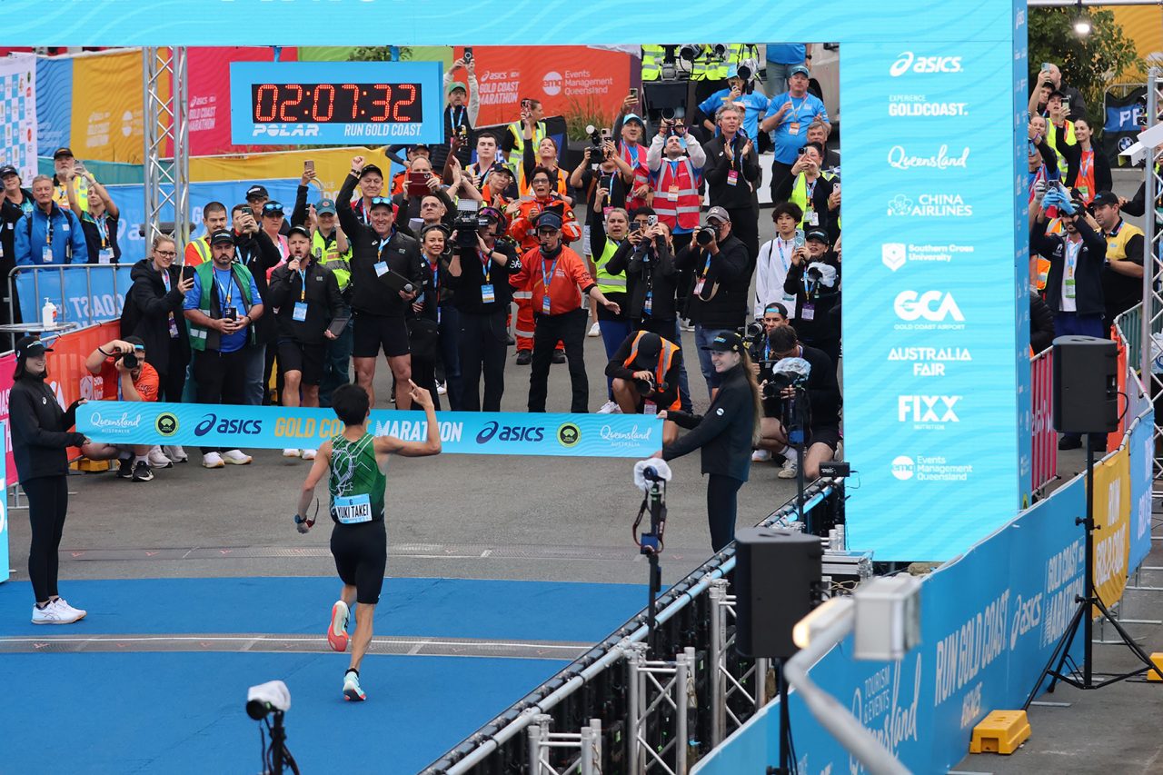 Runner crossing the finish area beside the Gold Coast Marathon display and cheering spectators.