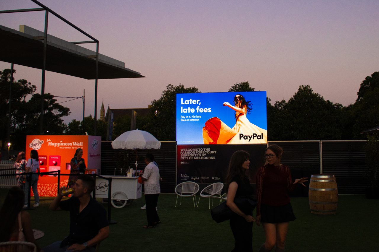 Outdoor digital advertising display beside food stalls at Melbourne Fashion Festival in the evening.