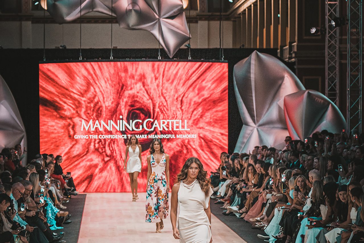 Manning Cartell branding on a red runway screen behind models at Melbourne Fashion Festival.
