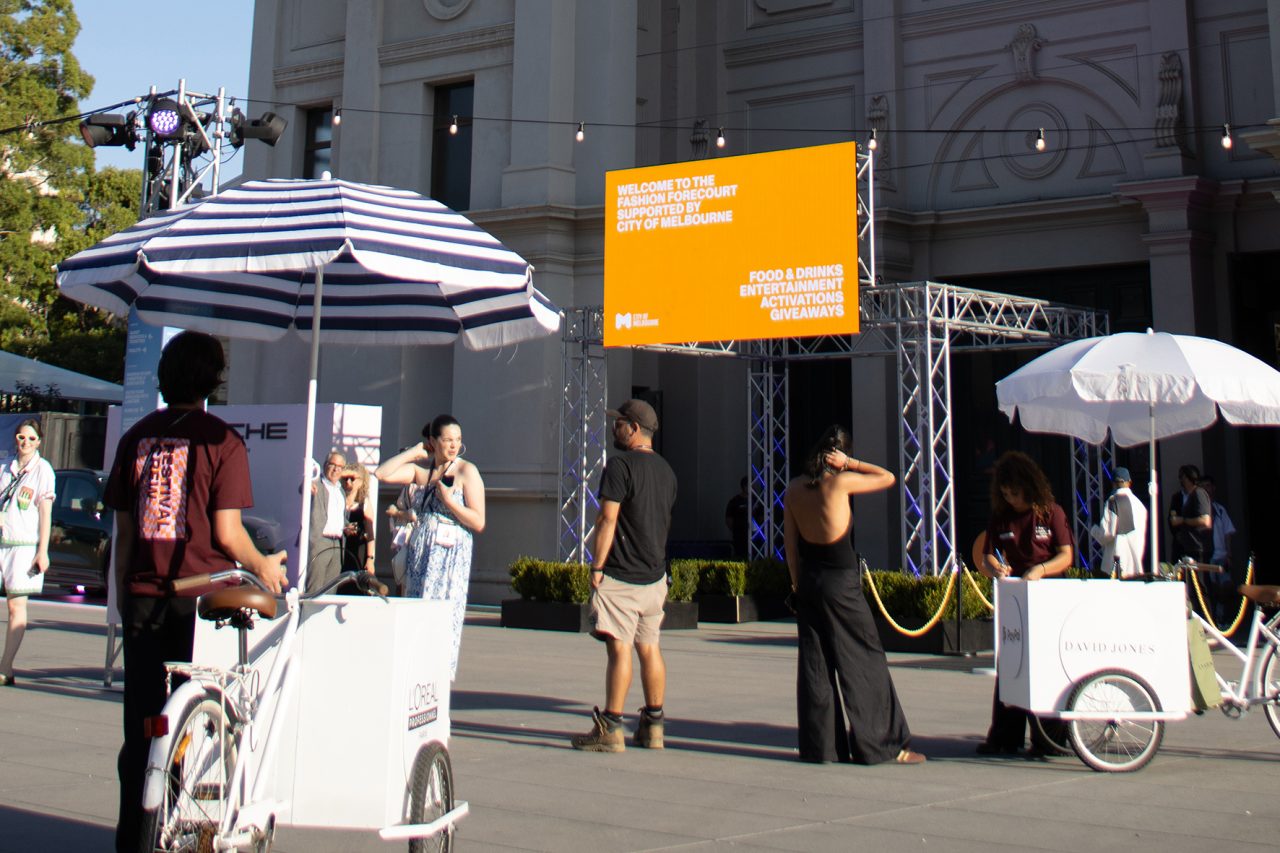 Orange digital screen above an outdoor activity area at Melbourne Fashion Festival 2025.
