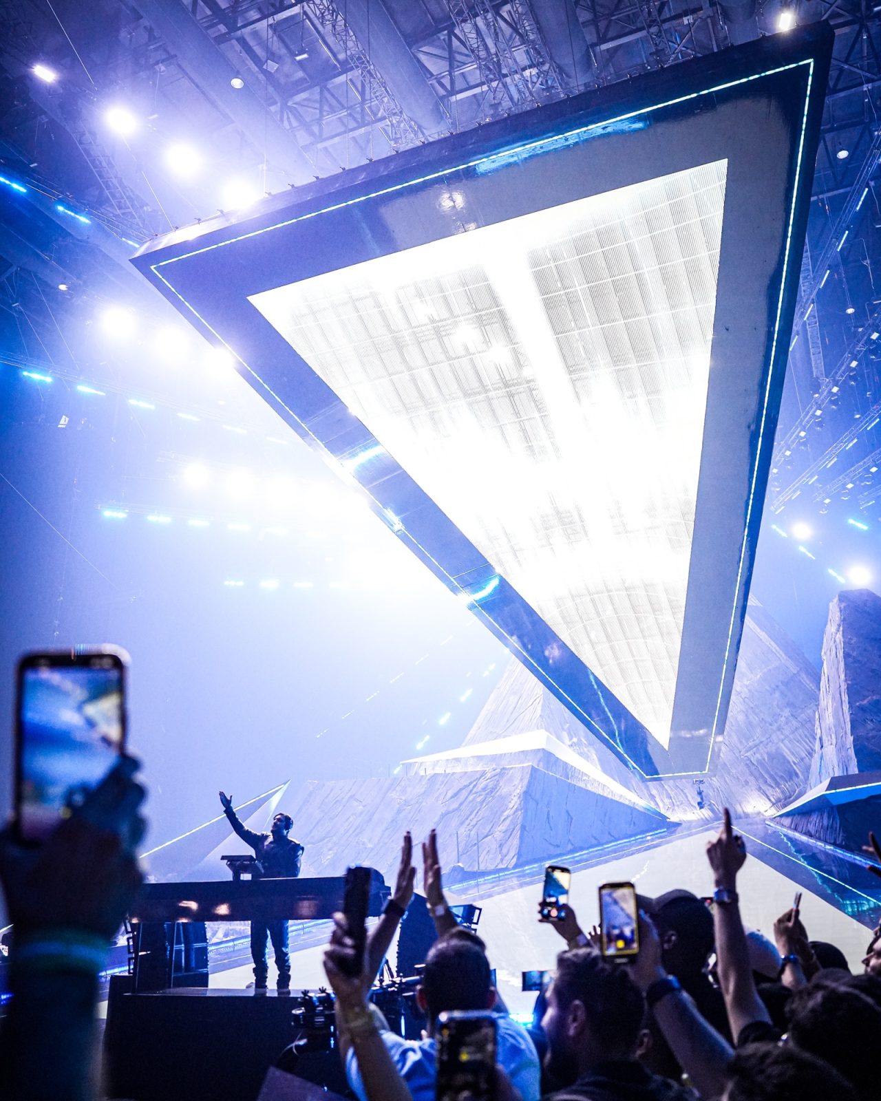 Large suspended triangular screen filled with white light above the audience.