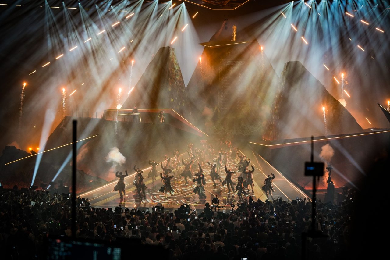 Performers beneath crossing white and amber spotlights at the opening ceremony.