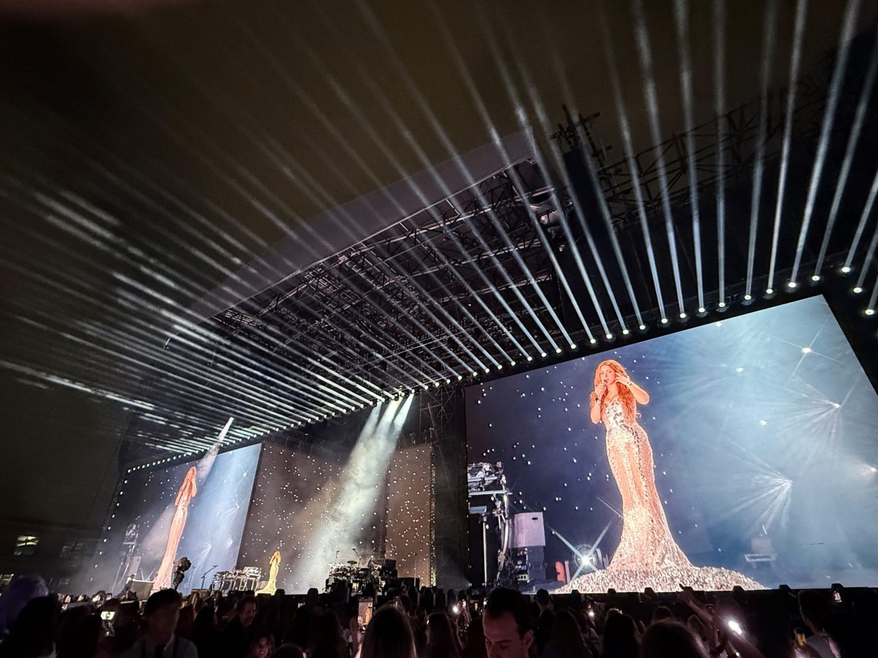 Shakira shown on a giant stage screen beneath parallel white beams.