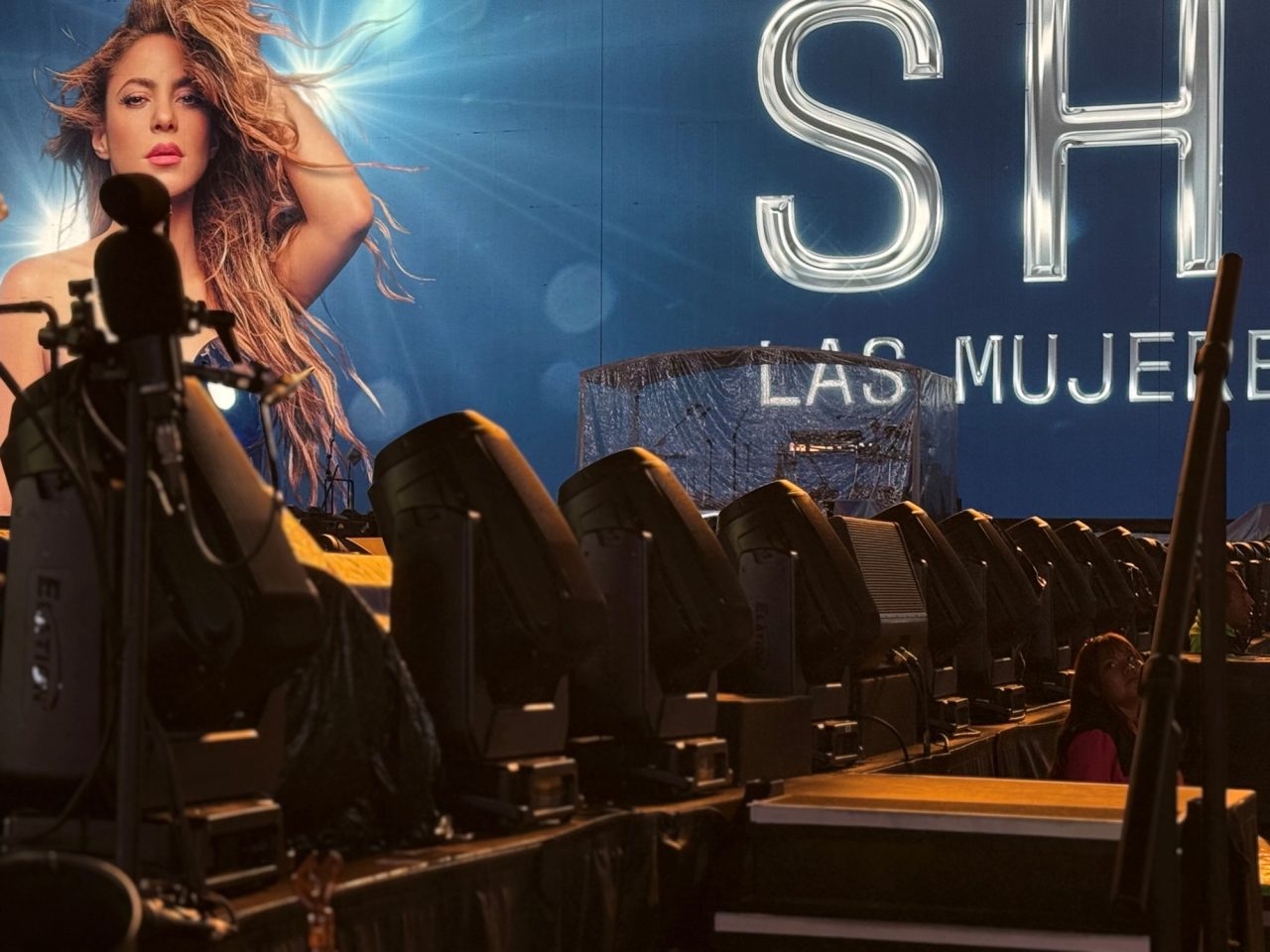 Row of lighting fixtures in front of a Shakira tour backdrop.