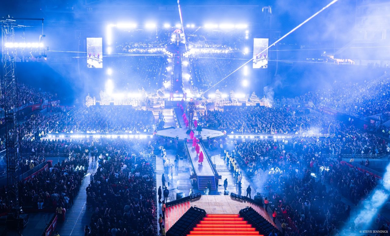 Long illuminated catwalk through a packed stadium under bright blue-white lighting.