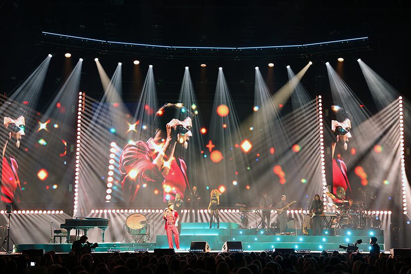 Live performance footage fills the video backdrop beneath white stage beams.