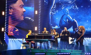 Guy Sebastian at a grand piano with backing vocalists, blue stage lighting and large video screens.