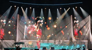 Guy Sebastian in a red suit beneath white beams, with his band and three tall video screens.